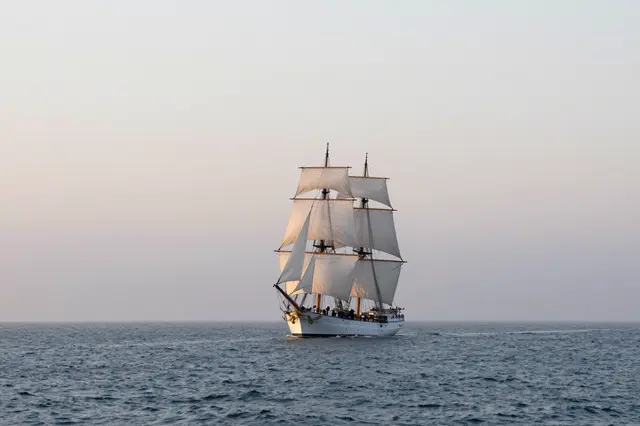 A tall sailing ship with billowing white sails glides across a misty calm sea at soft dawn