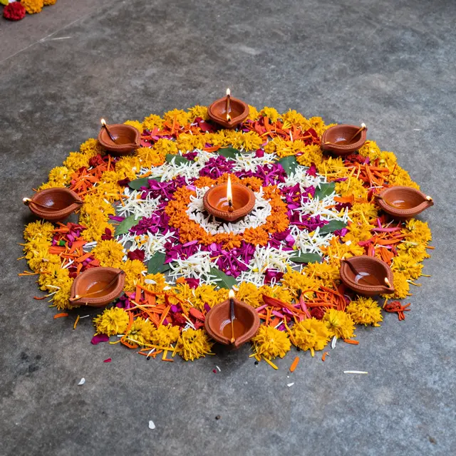 A circular rangoli of colorful flower petals sits with lit clay oil lamps on a grey stone floor for Diwali.