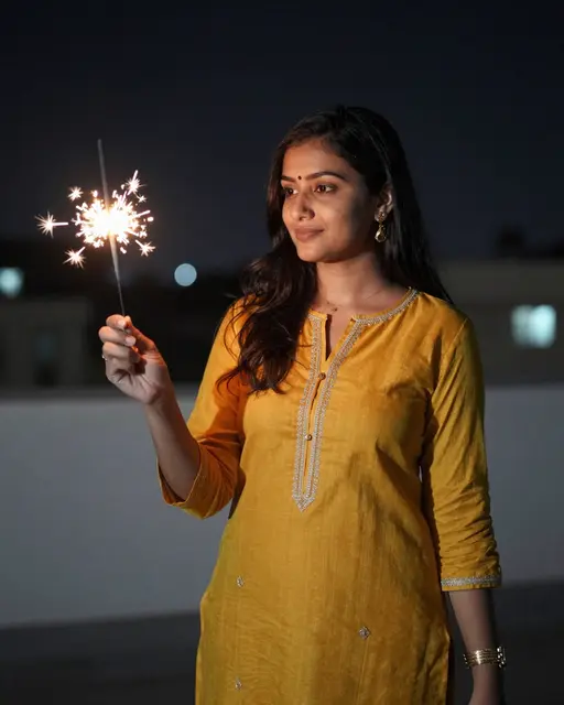 A woman in a mustard kurta holds a lit sparkler on a rooftop at night during Diwali