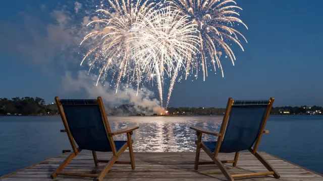 Two lawn chairs sit on a wooden dock facing bright fireworks bursting over a calm lake at dusk