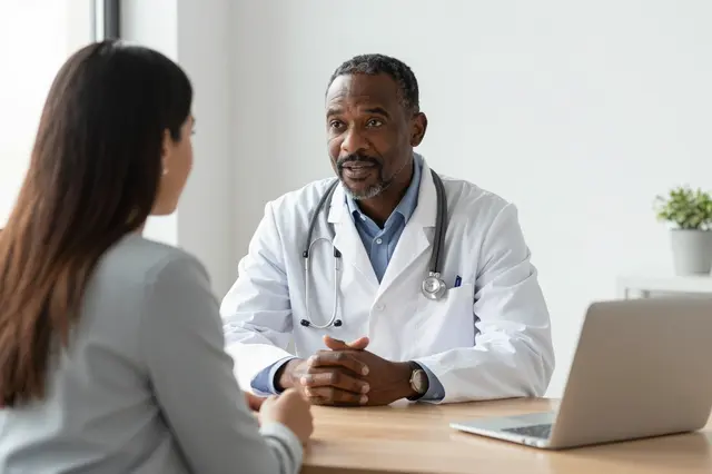 A middle aged Black doctor talks with a woman across a wooden desk in a bright medical office