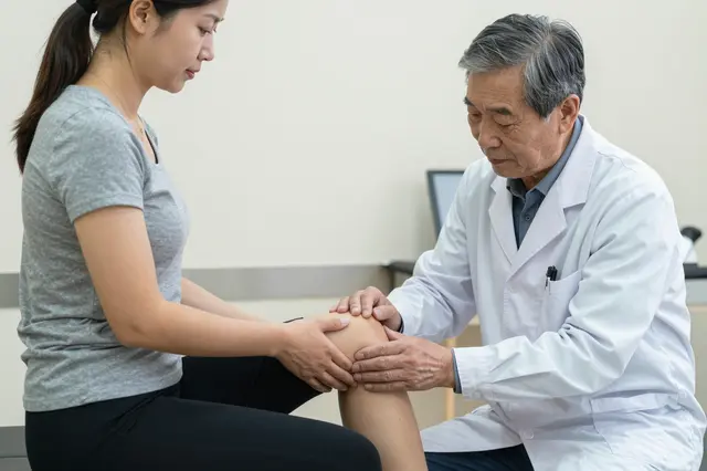 Senior East Asian doctor examines the knee of a young woman seated on a clinic exam table