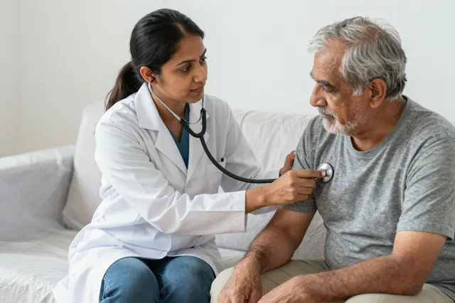 A South Asian female doctor uses a stethoscope to examine an older male patient seated on a light couch