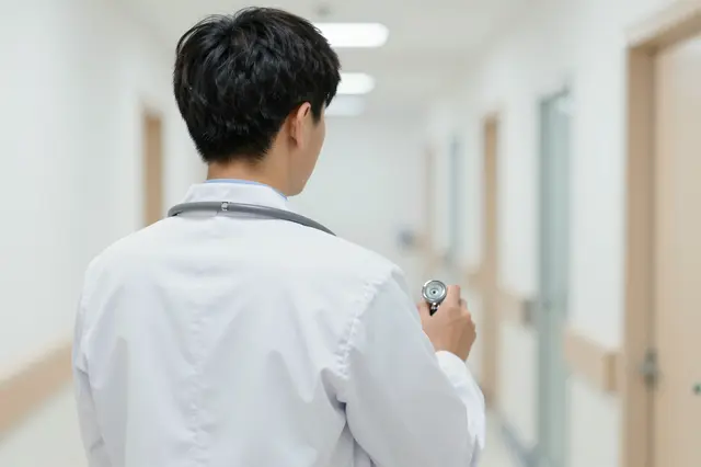 View from behind of a doctor wearing a lab coat and stethoscope standing in a bright hospital corridor.