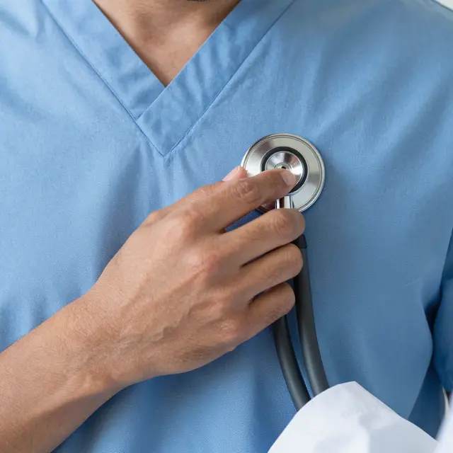 Close-up of a hand pressing a stethoscope to light blue medical scrubs worn by a person.