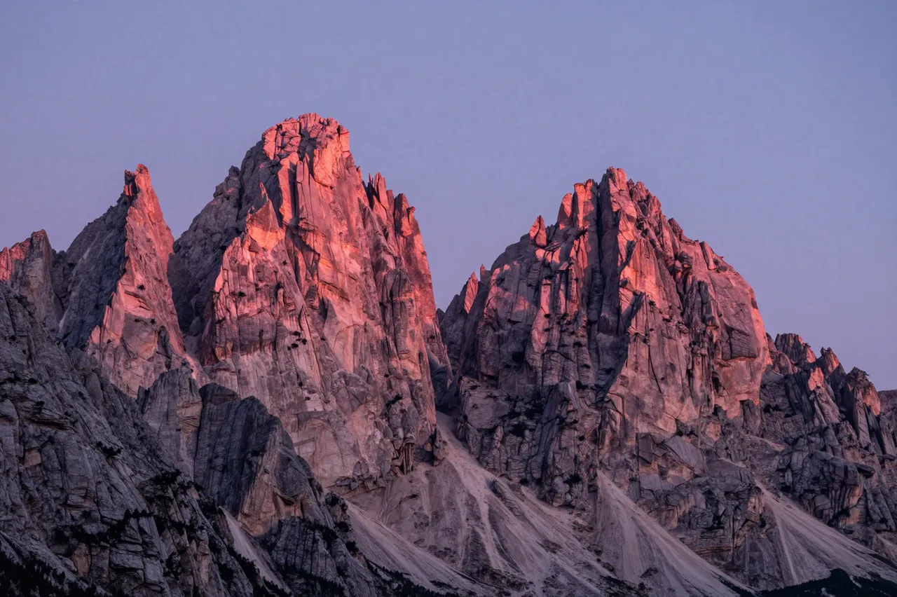 Jagged limestone Dolomite peaks glow with warm pink alpenglow against a pale twilight sky at sunset