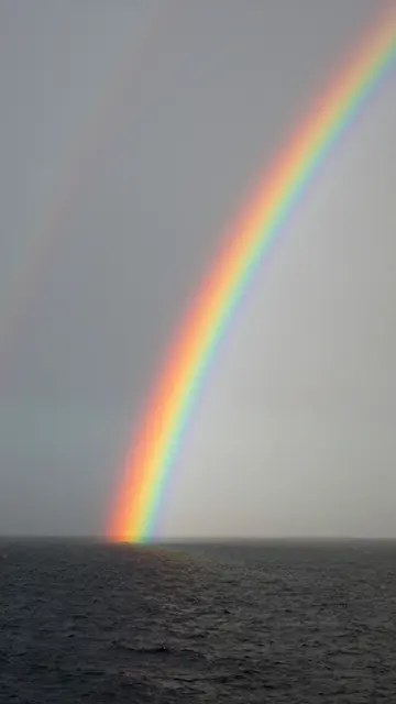 A bright primary rainbow with a faint secondary arc extends from a dark choppy ocean under an overcast gray sky
