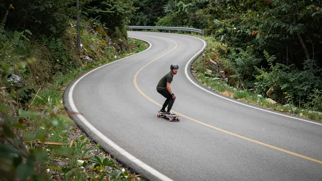 A helmeted adult man longboards down a winding forested mountain road