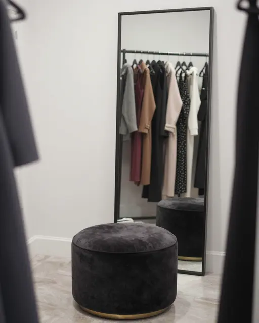 A modern dressing room features a dark velvet ottoman next to a tall framed floor mirror reflecting clothing on a rack.