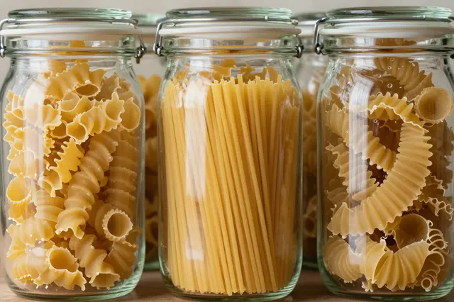 Sealed glass storage jars filled with assorted dry pasta varieties sit on a wooden surface