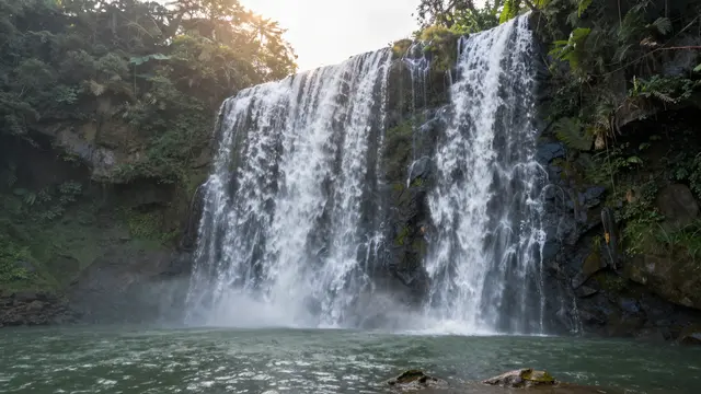 Two broad cascading waterfalls empty into a lush jungle pool as warm sunrise light filters through overhead trees