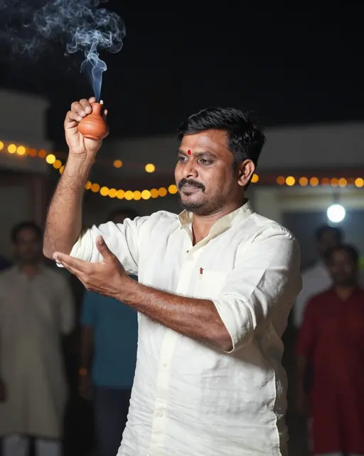 South Asian man holds a smoking clay incense burner during a nighttime festive gathering with warm string lights behind him