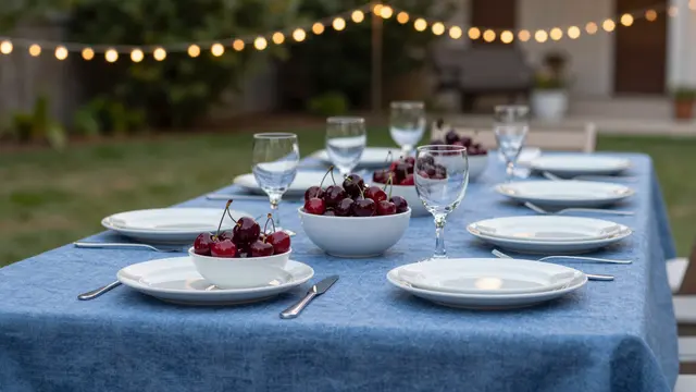 Long outdoor dining table with blue linen, place settings of white plates, cherry bowls and glasses under glowing string