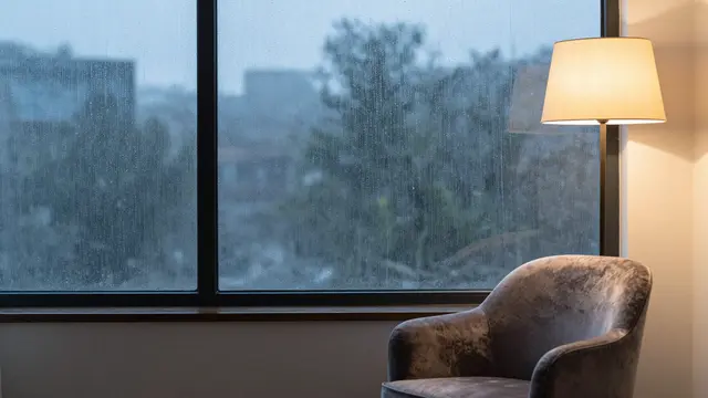 A plush velvet armchair sits beside a glowing floor lamp next to a rain-streaked window at dusk