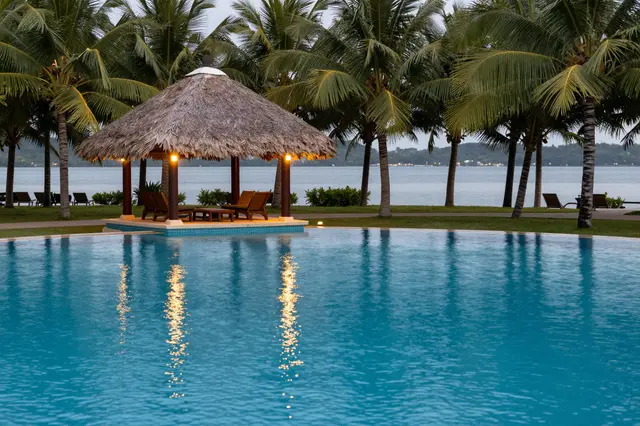 Lit thatched pool pavilion sits beside a calm turquoise resort pool lined with coconut palms at twilight