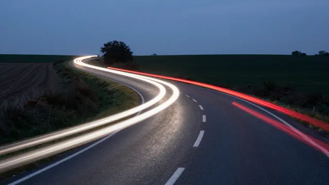 Long exposure red and white vehicle light trails curve along a winding rural country road at dusk