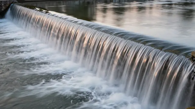 Smooth blurred water spills over a wide river weir captured with long exposure at dusk