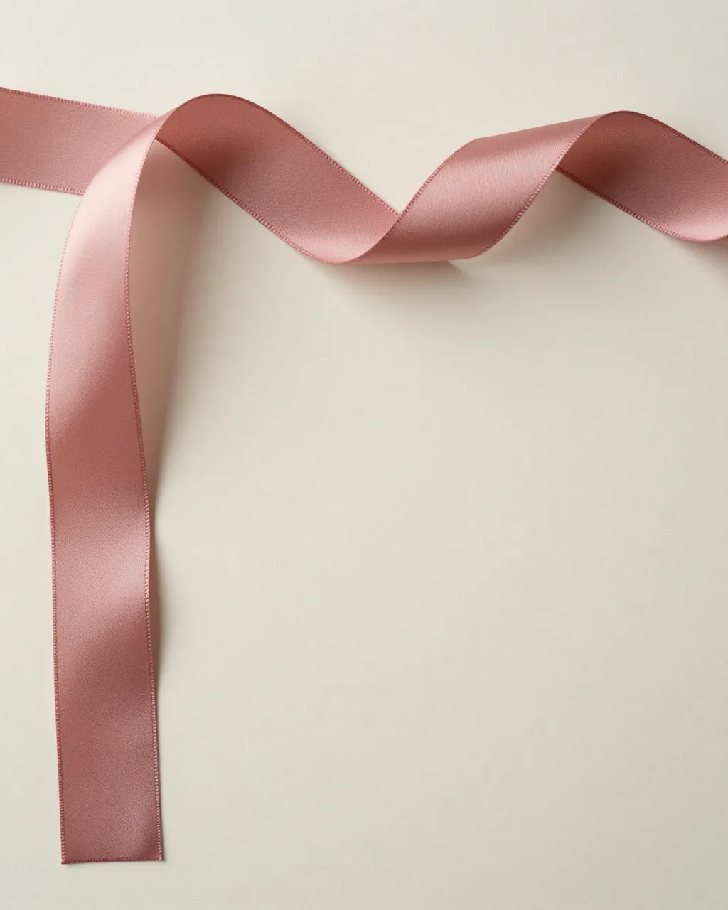 Curling dusty rose silk ribbon rests along the top edge of a smooth cream backdrop with empty copy space below.