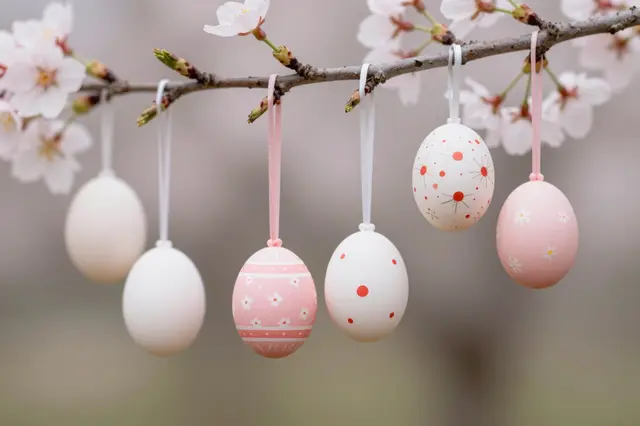 Decorated painted Easter eggs hang from a flowering cherry blossom branch against a soft blurred background