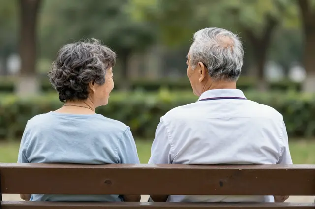Seen from behind, an elderly couple sits side by side on a wooden park bench looking out over green park grounds.
