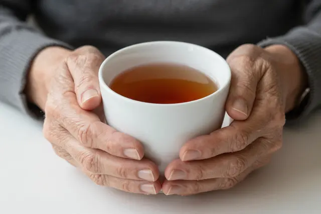Wrinkled elderly hands cradle a white ceramic cup filled with warm amber tea