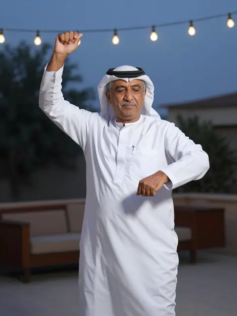 Senior Middle Eastern man dancing on an outdoor patio under string lights at twilight