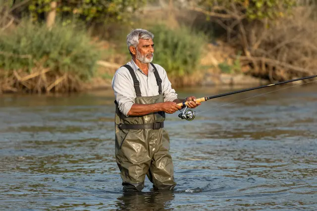 Senior South Asian man in chest waders stands in a shallow river holding a fishing rod at golden hour