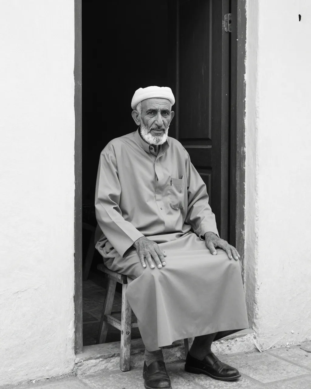 Black and white portrait of an elderly Middle Eastern man sitting on a wooden stool in a whitewashed doorway