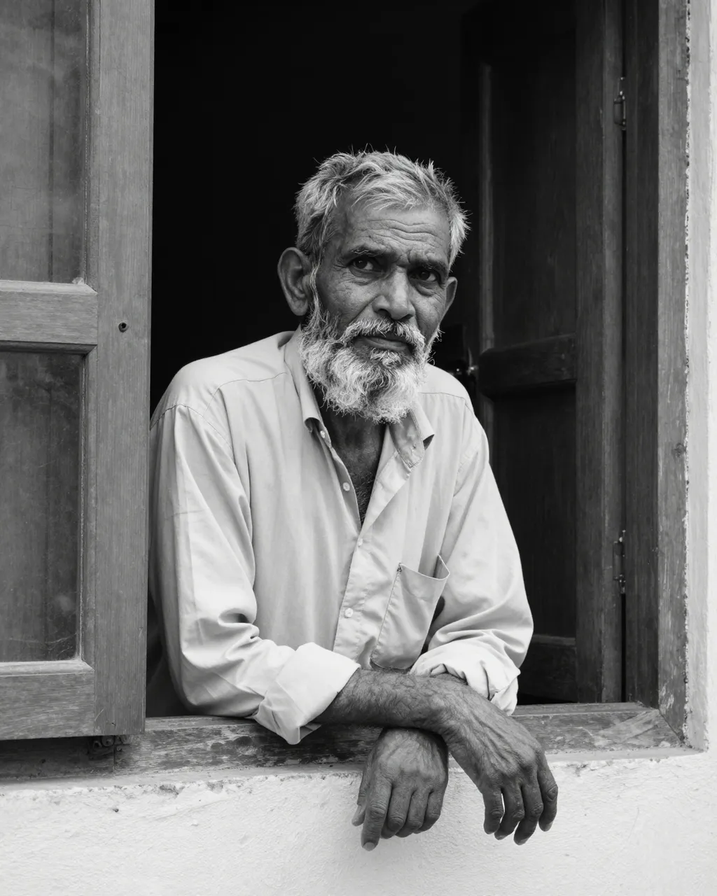 Black and white portrait of an elderly South Asian man with a gray beard leaning out of an open wooden window