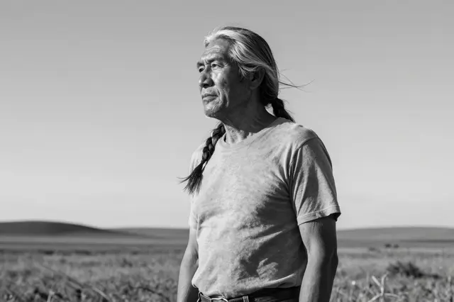 Black and white portrait of an older man with long gray hair gazing out over an open grassy plain
