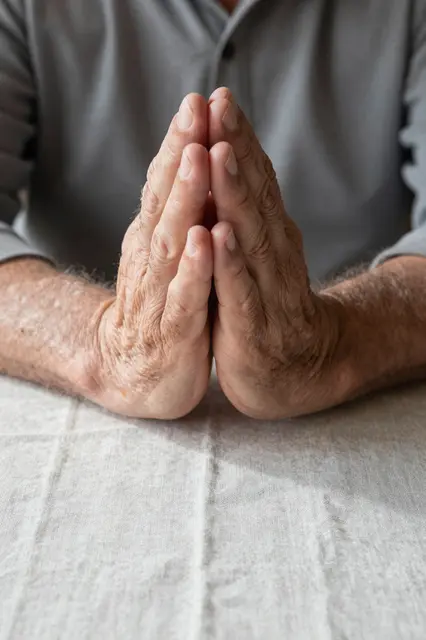 Weathered hands of an elderly man pressed together in prayer above a linen tablecloth in soft morning light