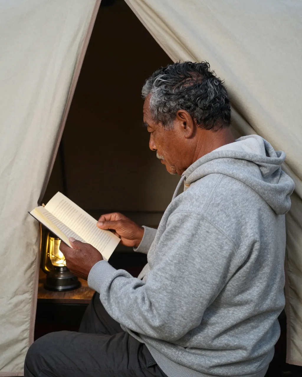 An older Pacific Islander man reads a book by lantern light while seated in a tent opening.