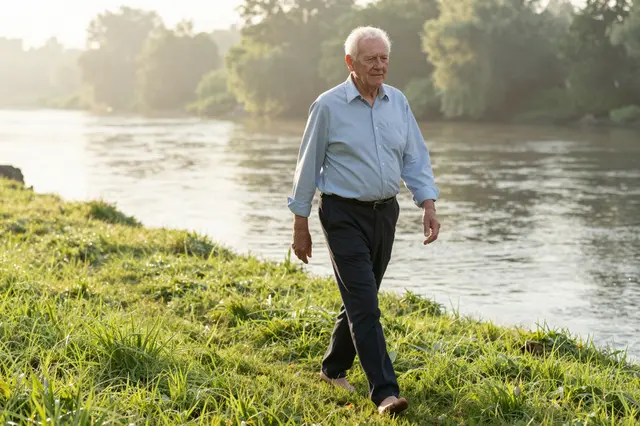 Barefoot elderly white man walks on dewy grass beside a misty river at dawn