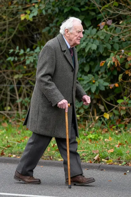 Side view of an elderly man in a tweed coat walking with a wooden cane beside leafy roadside vegetation