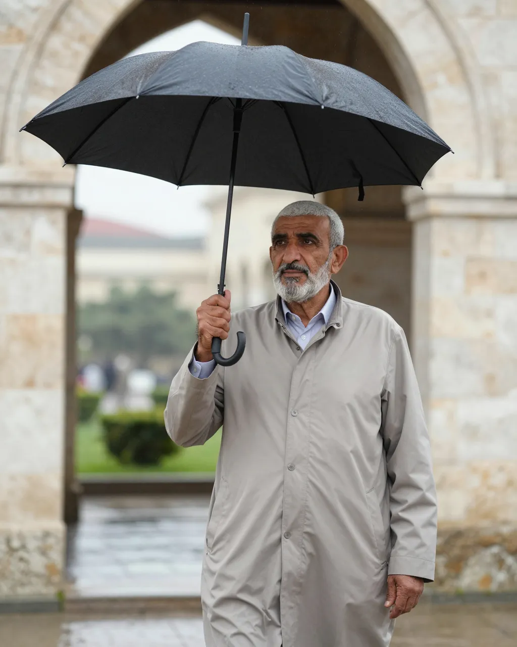 An older Middle Eastern man holds a black umbrella while standing under a stone arch on a rainy day.