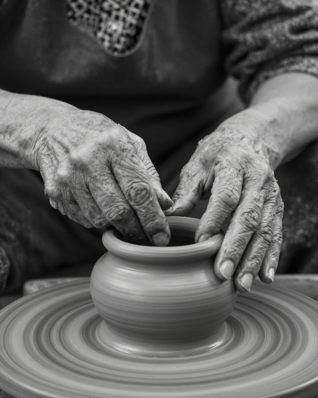 Close-up black and white view of weathered elderly hands shaping a clay pot on a spinning pottery wheel