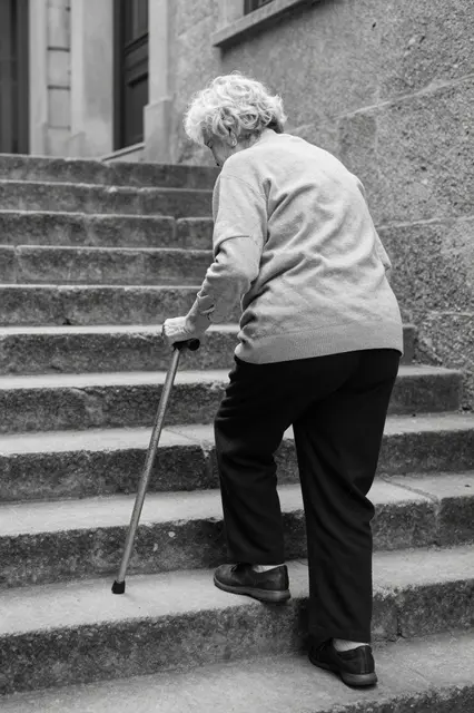 Back view of an elderly woman using a cane to climb exterior stone building stairs in black and white
