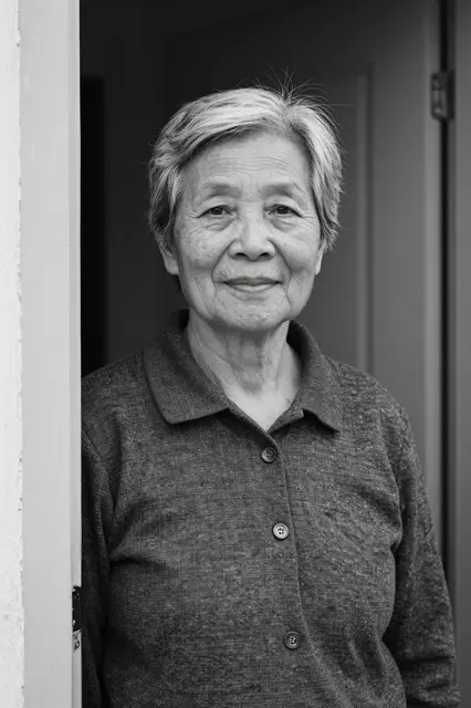 Black and white portrait of a senior East Asian woman with short gray hair standing in a doorway with a quiet smile