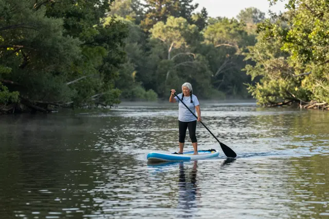 An elderly woman paddleboards on a calm forested river at misty dawn