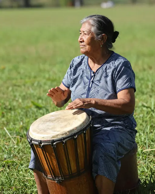 Side view of a senior Indigenous woman playing a wooden hand drum in a sunlit green meadow