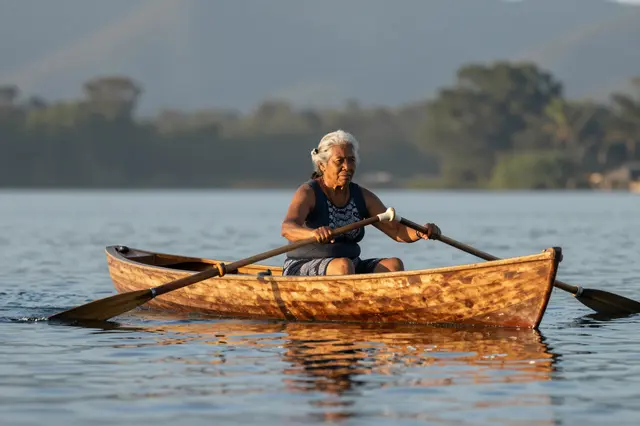 Senior Pacific Islander woman rows a wooden rowboat across calm still lake water at sunrise.