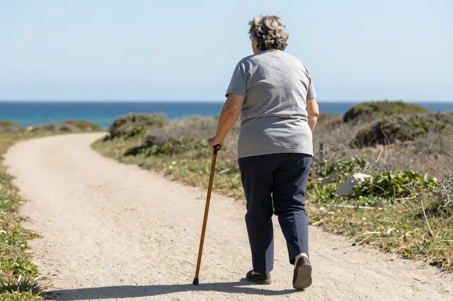 Back view of an elderly woman using a cane walking along a sunny dirt coastal path toward the ocean