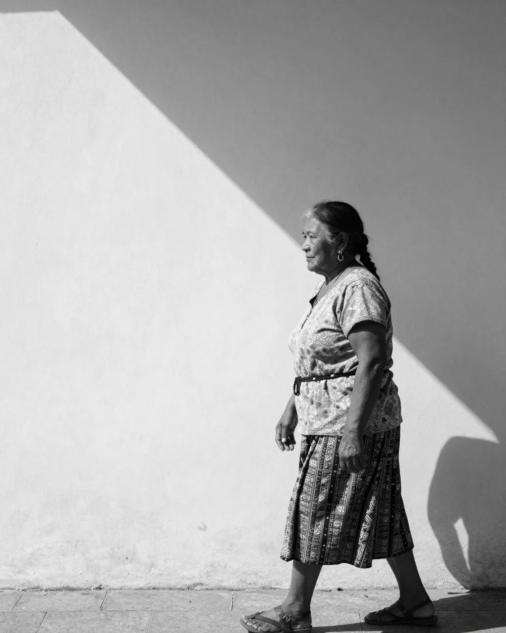 Black and white profile view of an older Indigenous woman walking along a sidewalk beside a wall split by a sharp diagonal