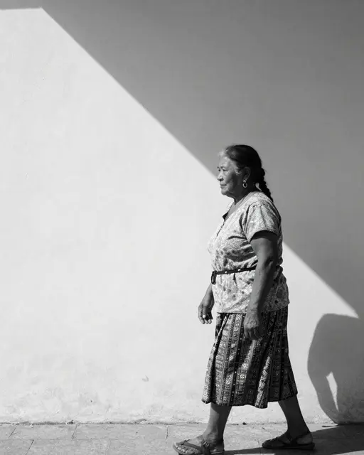 Black and white profile view of an older Indigenous woman walking along a sidewalk beside a wall split by a sharp diagonal