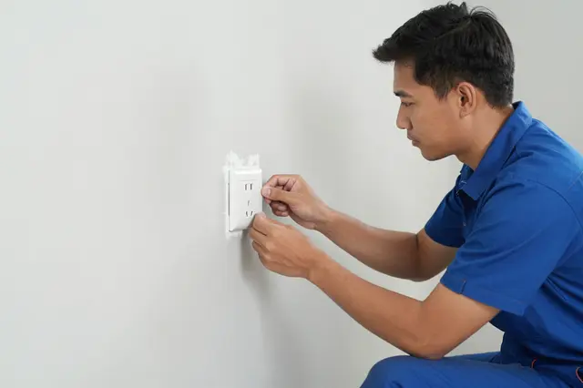A Southeast Asian electrician in a blue uniform fits a wall socket on a freshly plastered interior wall in side view.