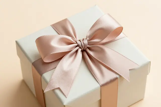 A square off-white gift box tied with a large blush satin bow rests on a soft peach background