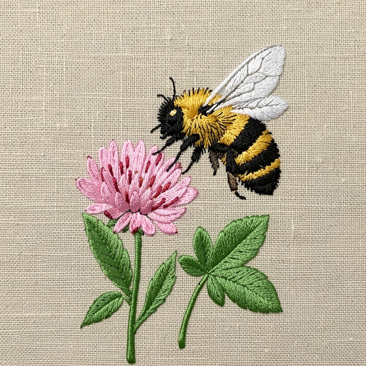 Fuzzy satin stitch embroidery of a bumblebee hovering near a pink clover flower on natural linen