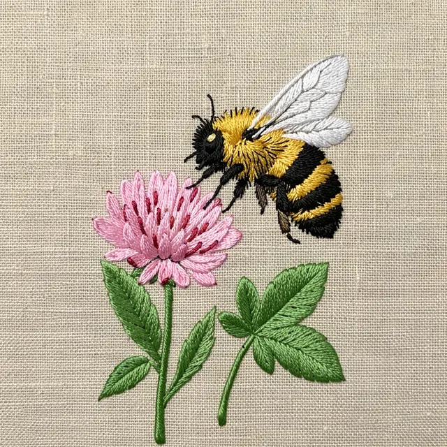 Fuzzy satin stitch embroidery of a bumblebee hovering near a pink clover flower on natural linen