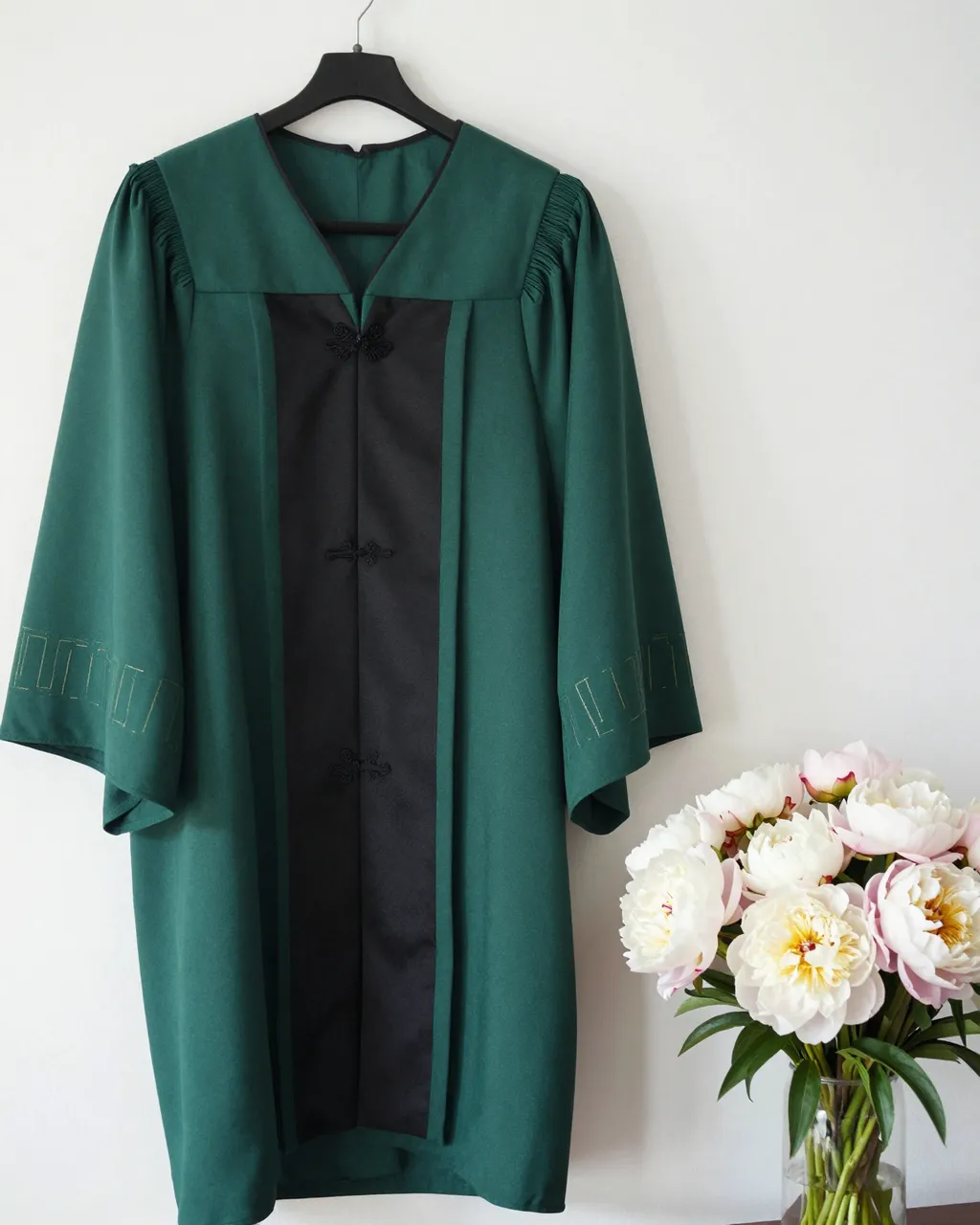 Green graduation gown hangs on a black hanger next to a peony bouquet against a plain white wall
