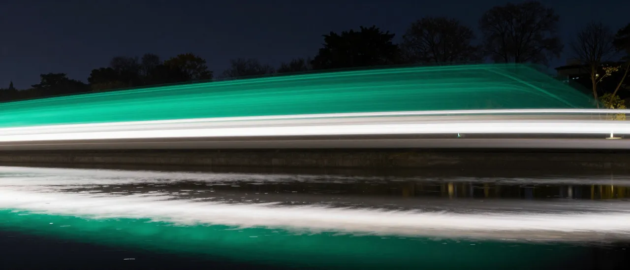 Long-exposure emerald and white light trails stretch across a night scene, mirrored on still water below